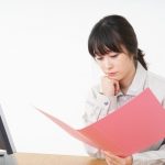 Woman reviews a pink folder while seated at a desk, looking at documents in front of a computer screen.