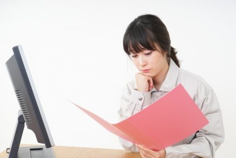 Woman reviews a pink folder while seated at a desk, looking at documents in front of a computer screen.